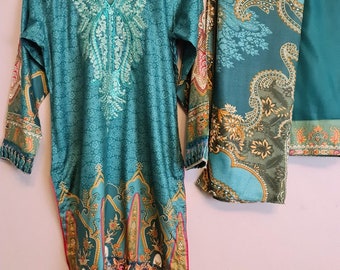 Op de afbeelding: Een tealblauwe lange jurk met lange mouwen met een paisley print en gouden borduurwerk. De jurk wordt gecombineerd met een bijpassende tealblauwe sjaal met een paisley print.