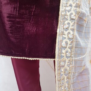 Puede incluir: Una blusa de terciopelo granate con ribete bordado blanco y dorado. La blusa se combina con pantalones granates.