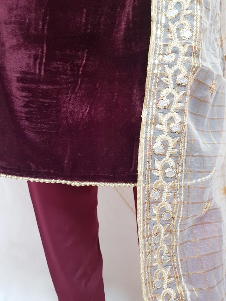 Puede incluir: Una blusa de terciopelo granate con ribete bordado blanco y dorado. La blusa se combina con pantalones granates.