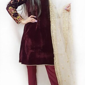 Puede incluir: Una kurta de terciopelo granate con bordados dorados y un pantal&oacute;n a juego. La kurta tiene mangas largas y un escote redondo. El atuendo se combina con una dupatta blanca transparente con bordados dorados.