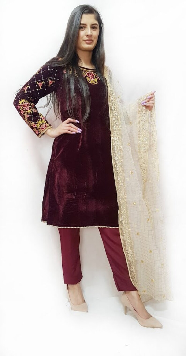 Puede incluir: Una kurta de terciopelo granate con bordados dorados y un pantal&oacute;n a juego. La kurta tiene mangas largas y un escote redondo. El atuendo se combina con una dupatta blanca transparente con bordados dorados.