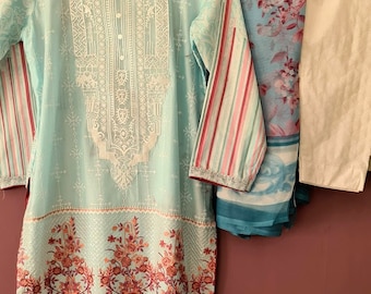 Op de afbeelding: Een lichtblauwe, langarmige, geborduurde jurk met een bloemenprint en een bijpassende dupatta. De dupatta is een lichtblauwe, doorschijnende stof met een bloemenprint. De jurk heeft een witte geborduurde halslijn en een witte geborduurde zoom. De jurk hangt aan een houten hanger.