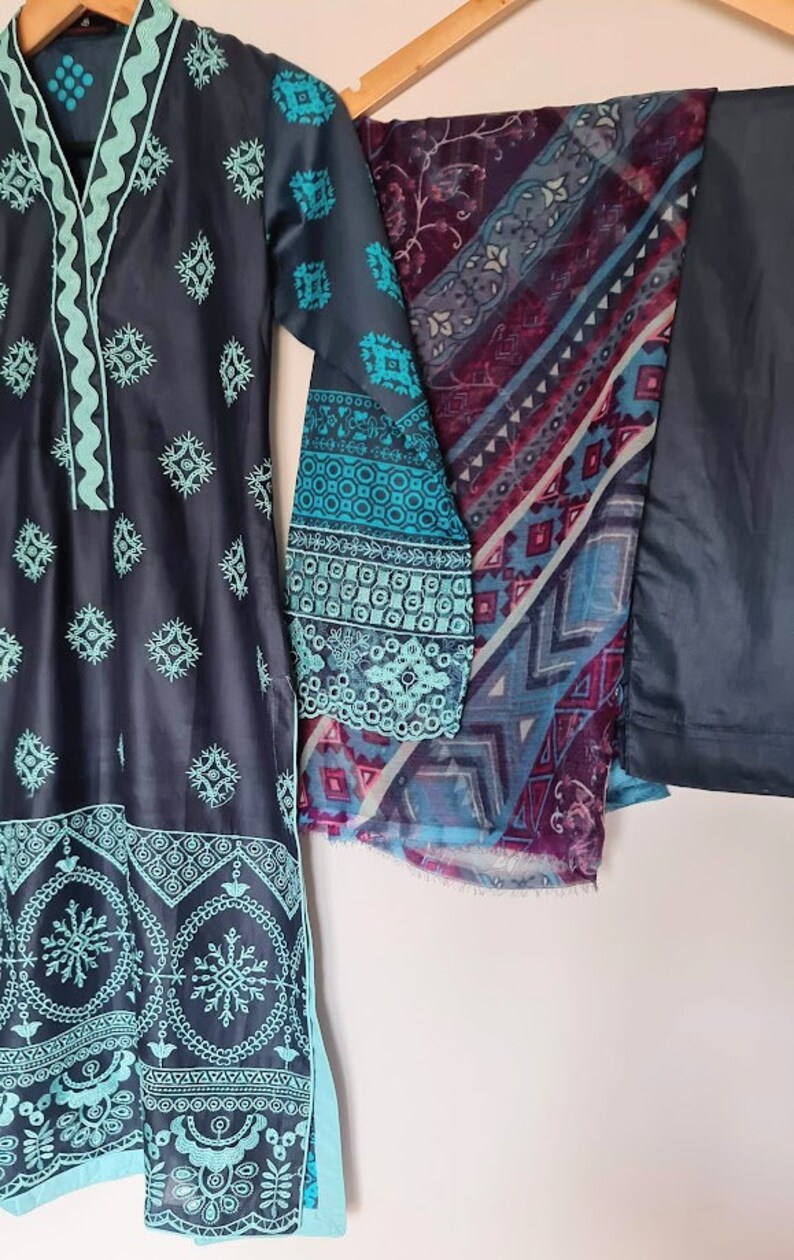 May include: A dark blue and teal embroidered dress with a matching dupatta and pants. The dress has a V-neckline and a floral pattern. The dupatta has a geometric pattern. The pants are solid dark blue.