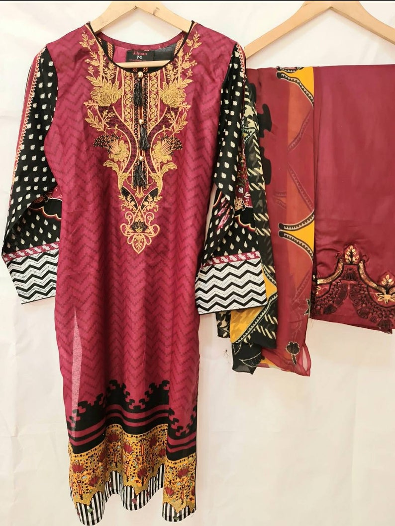 May include: A maroon and black printed three-piece dress set with a long-sleeved top, a matching bottom, and a dupatta. The top features a floral design with gold embroidery and a tassel. The bottom has a chevron pattern and the dupatta has a floral design with gold accents.