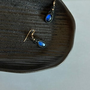 May include: A pair of earrings with blue gemstone accents and black macrame detailing. The earrings are hanging from a black surface.