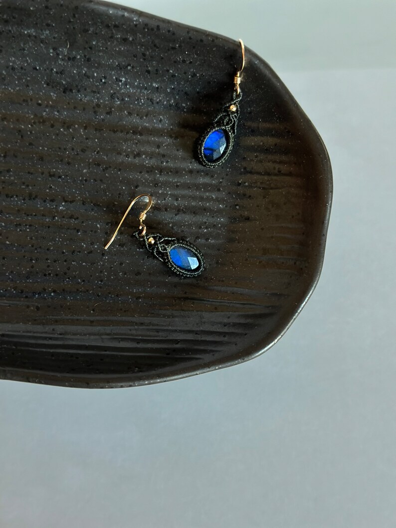May include: A pair of earrings with blue gemstone accents and black macrame detailing. The earrings are hanging from a black surface.