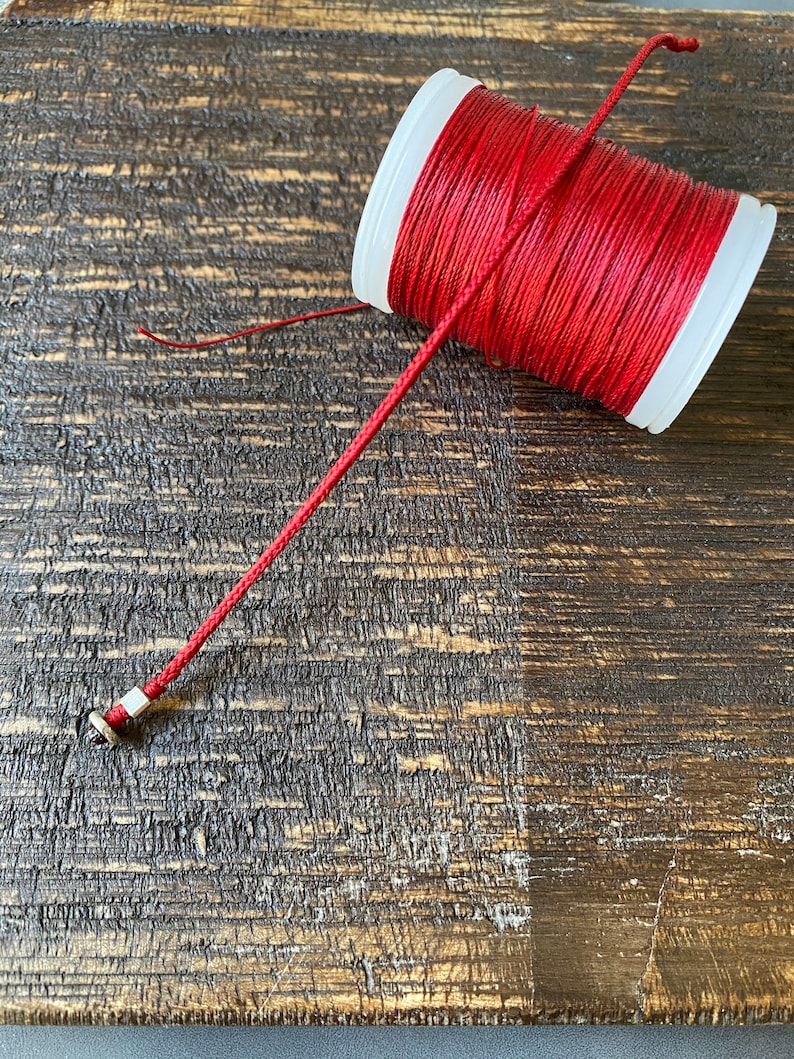 May include: A spool of red waxed cord on a wooden surface. The cord is wound around a white plastic spool.