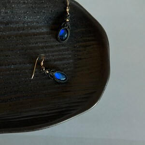 May include: A pair of dangle earrings with blue gemstone accents and black thread wrapped around the metal. The earrings are on a black plate.
