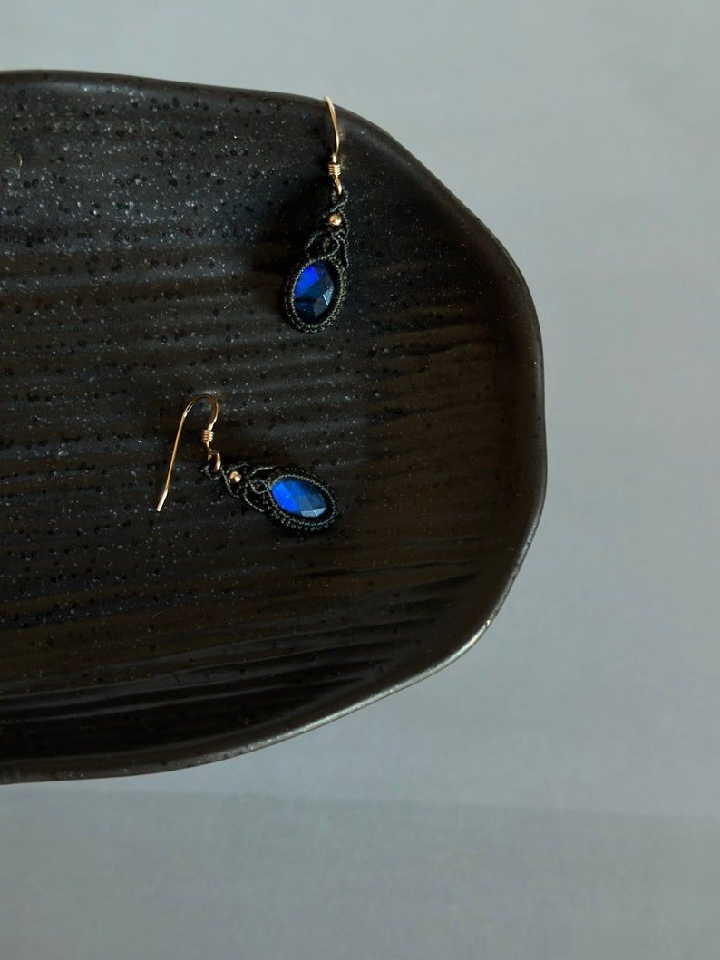 May include: A pair of dangle earrings with blue gemstone accents and black thread wrapped around the metal. The earrings are on a black plate.