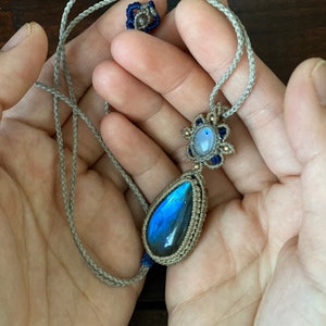 May include: A blue labradorite gemstone pendant necklace with a smaller moonstone gemstone in a macrame knot design. The necklace is made with a beige cord.