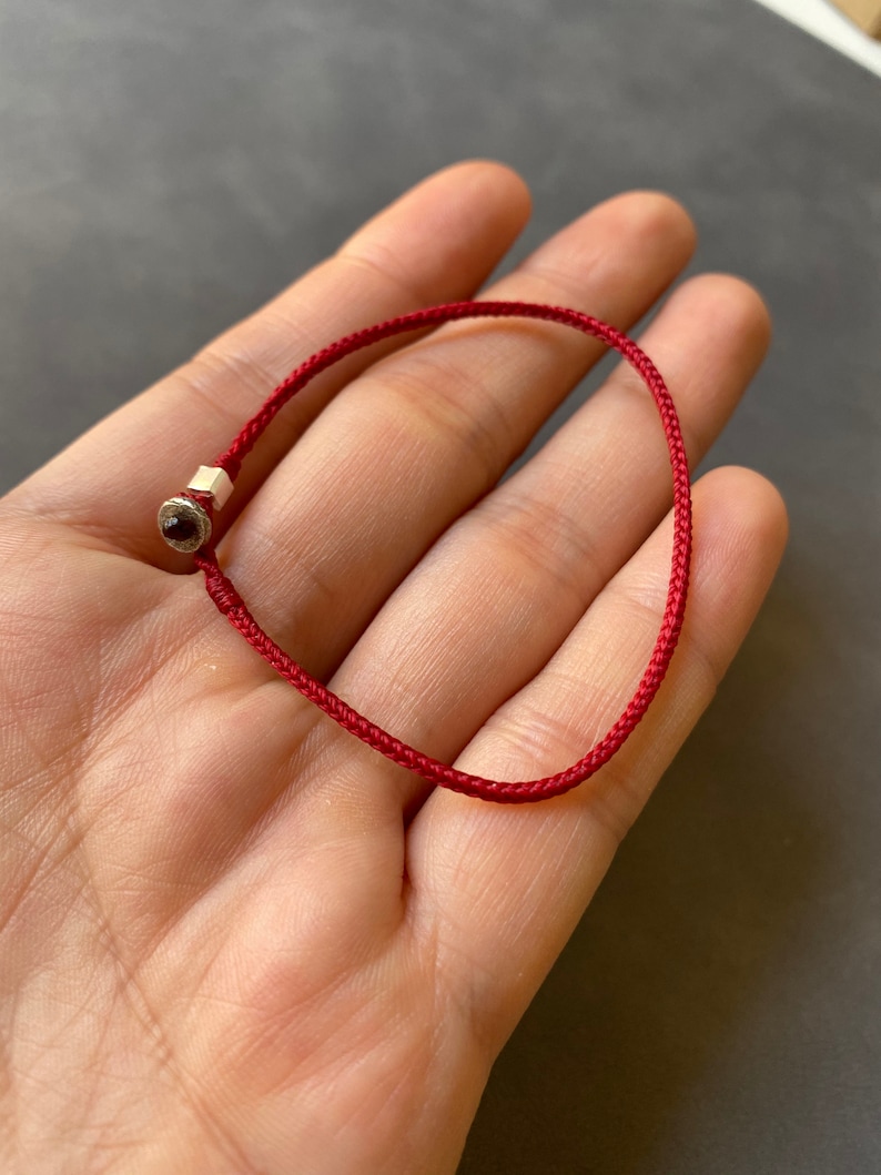 May include: A red string bracelet with a small silver charm featuring a red gemstone.