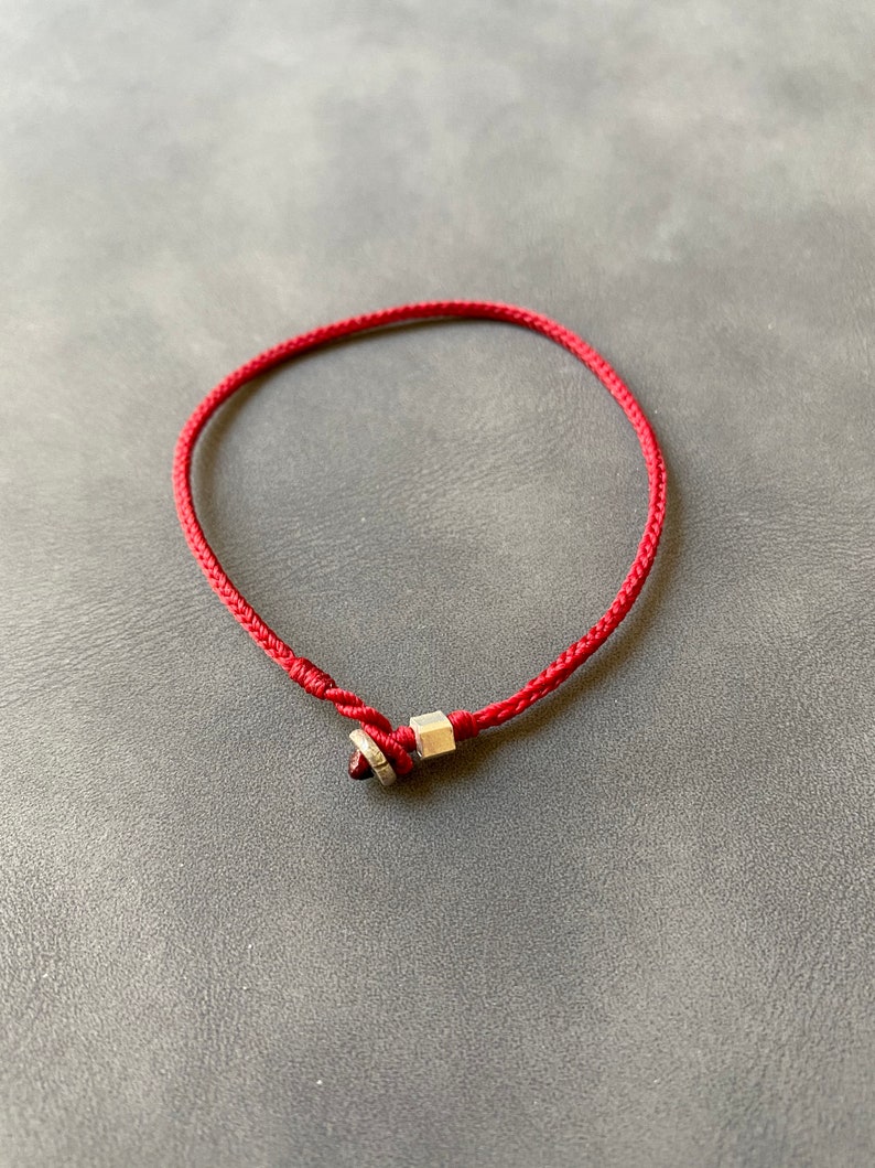 May include: A red string bracelet with a silver charm. The charm has a small red stone in the center.