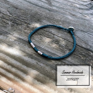 May include: Teal braided bracelet with silver-toned accents. The bracelet features a woven design and a circular clasp. The bracelet is displayed on a weathered wooden surface. The text "Summer Handmade 20190217" is in a white box.