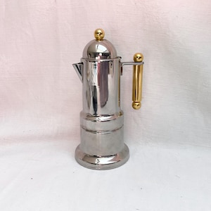 May include: A polished silver-tone metal stovetop coffee maker featuring a cylindrical body, a small spout, and a vertical handle. The lid is topped with a small gold-tone sphere, and the handle is accented with matching gold-tone cylindrical details.