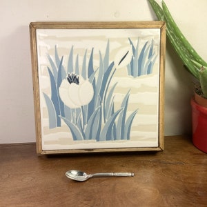 May include: A framed ceramic tile with a blue and white floral design. The tile features stylized white flowers with blue leaves on a cream background.