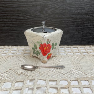 Peut inclure: Un sucrier en céramique blanche avec un couvercle argenté et un motif floral avec des roses rouges. Le bol est posé sur une dentelle blanche crochetée avec une cuillère en argent à côté.