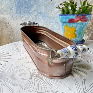 May include: An oval copper basin featuring two side handles made of white ceramic with a blue floral pattern. The basin has a metallic finish and a smooth, rounded rim. The handles are attached to the copper body with curved metal brackets.