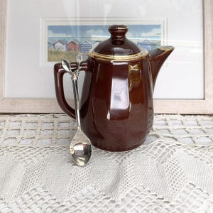 May include: A glossy brown ceramic teapot with a gold trim and lid, accompanied by a silver spoon. The teapot rests on a white lace tablecloth. A framed picture of beach huts is in the background.