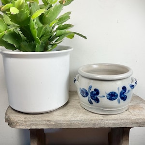 May include: Two ceramic plant pots on a wooden surface. A large white pot contains a green plant. A smaller, light-coloured pot with blue floral designs and handles sits beside it. The pots are suitable for indoor or outdoor use.