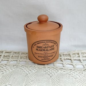 May include: A tan ceramic jar with a lid, labeled "Breakfast Marmalade." The jar has a round body and a small knob on the lid. The text on the jar reads "The Original Suffolk Jar" and "Henry Watson Pottery, Founded 1800."