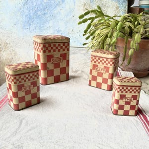 Peut inclure: Quatre boîtes de rangement vintage en métal avec des motifs à carreaux rouges et crème. Les boîtes sont étiquetées "SUCRE", "CHICOREE", "CAFE" et "THE". Les boîtes sont carrées avec un couvercle assorti.