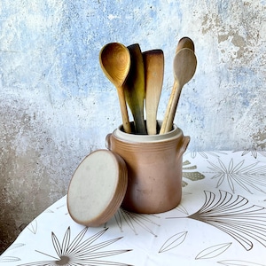 Puede incluir: Un conjunto de utensilios de cocina de madera, incluyendo cucharas y espátulas, se exhiben en una olla de cerámica marrón con tapa. La olla está sobre un mantel blanco con un patrón de hojas, contra una pared texturizada.