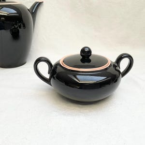 May include: A black ceramic tea set with a teapot and a sugar bowl. The teapot has a long spout and a curved handle. The sugar bowl has two handles and a lid with a small knob. Both pieces have a glossy finish and a pink accent.