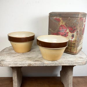 Op de afbeelding: Twee vintage keramische kommen met een bruine rand en een lichtgele body. Een rechthoekige metalen doos met een vervaagd rood en wit bloemenontwerp staat achter de kommen. De items worden tentoongesteld op een verweerde houten kruk.