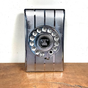 Pode incluir: Um telefone de parede retangular com acabamento cromado e um disco giratório circular. O disco inclui botões circulares brancos rotulados com pares de letras de AB a YZ e um ícone central preto representando um auscultador de telefone clássico.