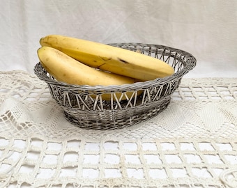Puede incluir: Una cesta de alambre plateado contiene tres plátanos maduros. La cesta ovalada tiene un diseño tejido y descansa sobre un mantel de encaje blanco. Los plátanos son amarillos con algunas manchas marrones.