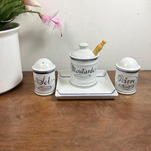 Peut inclure: Un ensemble de condiments en céramique blanche avec des bordures bleues, comprenant un salière, un poivrier et un pot à moutarde avec une cuillère en bois. L'ensemble repose sur un plateau rectangulaire assorti. Les mots "Sel", "Moutarde" et "Poivre" sont écrits.
