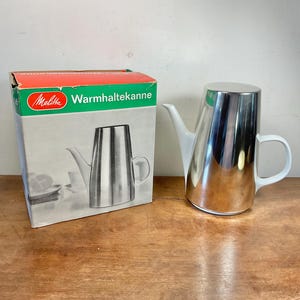 Peut inclure: Une cafetière Melitta Warmhaltekanne en acier inoxydable avec une poignée et un bec verseur blancs, à côté de sa boîte d'origine. La boîte a un dessus rouge et vert avec le logo Melitta et le nom du produit.