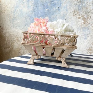 Puede incluir: Una cesta rectangular de metal color blanco roto con cuatro patas ornamentadas y curvadas, y un borde decorativo calado. La cesta está llena de un grupo de pétalos de flores artificiales suaves en tonos rosa pálido y blanco, formando un conjunto.