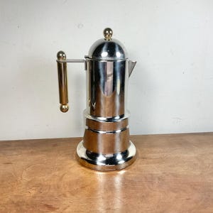 Peut inclure: Une cafetière italienne en métal argenté brillant avec un corps cylindrique, un couvercle bombé surmonté d'un petit bouton sphérique et une poignée verticale avec des accents dorés en haut et en bas. Elle possède un petit bec verseur.