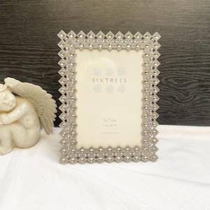 May include: A decorative silver-toned picture frame with a geometric design, holding a 5x7 inch photo. The frame is next to a small, off-white cherub statue with folded wings. The frame has the text "SIXTREES".