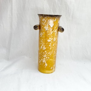 Pode incluir: Um vaso cilíndrico com uma cor base amarelo-mostarda e um padrão salpicado de branco por toda a superfície. O vaso tem uma borda alargada e duas pequenas alças castanho-escuras em forma de voluta nos lados superiores do corpo.