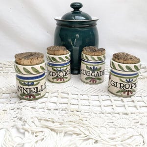Puede incluir: Un conjunto de botes de especias de cerámica con tapones de corcho y un gran bote de cerámica verde. Los botes pequeños están etiquetados con nombres de especias franceses y decorados con hojas verdes, rayas azules y detalles florales rojos. El bote grande tiene tapa.
