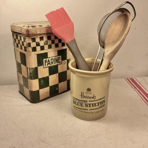 Könnte beinhalten: Ein Keramik-Utensilienhalter mit goldener Glasur und schwarzem Text, der "Harrods Finest Selected Blue Stilton Cheese" lautet. Im Halter befinden sich eine rote Silikon-Backbürste, ein Holzlöffel und ein silbernes Drahtsieb.