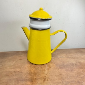 May include: A vintage yellow enamel coffee pot with a white and black trim. The pot has a lid and a curved handle. The coffee pot is on a wooden surface.