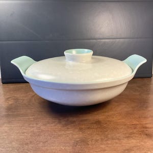 Peut inclure: Un plat à casserole vintage en céramique blanc cassé avec un couvercle. Le plat présente un motif moucheté et des accents bleu clair sur les poignées et le bouton du couvercle. Le plat est posé sur une surface en bois.