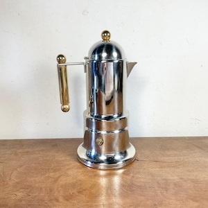 May include: A polished chrome stovetop coffee maker featuring a cylindrical body, a domed lid with a small gold-toned spherical knob, and a vertical side handle with gold-toned accents at the top and bottom, including a small gold-toned valve.