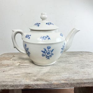 May include: A white ceramic teapot with a lid, handle, and spout. The teapot is decorated with blue floral patterns. The teapot is sitting on a wooden surface.