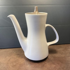 Può includere: Una caffettiera in ceramica bianca con un coperchio marrone. La caffettiera ha un corpo arrotondato e un beccuccio laterale. Il coperchio ha un piccolo pomo in cima.