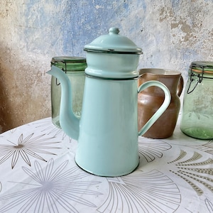 Puede incluir: Una cafetera vintage de esmalte azul turquesa con tapa, boquilla y asa. Junto a ella, una jarra de cerámica marrón y dos tarros de cristal con tapas. Los objetos están sobre un mantel blanco con estampado floral.