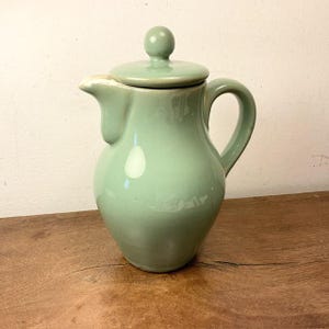 May include: A light green ceramic creamer with a lid and a handle. The creamer has a rounded body and a smooth, glossy finish.
