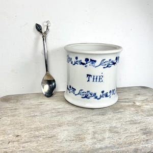 Puede incluir: Un tarro de té de cerámica blanca con diseños florales azules y la palabra "THE" en azul. Una cuchara plateada con un detalle de cristal descansa junto al tarro. Los artículos están sobre una superficie de madera envejecida.