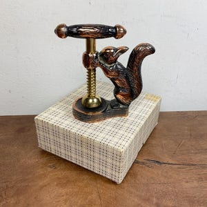 Puede incluir: Un cascanueces de metal con una figura de ardilla en acabado bronce oscuro y un mecanismo de tornillo roscado en tono dorado. El cascanueces está montado sobre una base rectangular y colocado sobre una caja blanca con un patrón de rejilla negra.