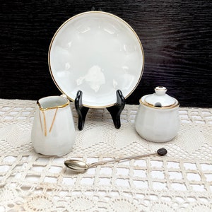 May include: White porcelain creamer, sugar bowl, and plate with gold trim. The creamer and sugar bowl have a gold design on the front. A silver spoon is on a white crocheted tablecloth.