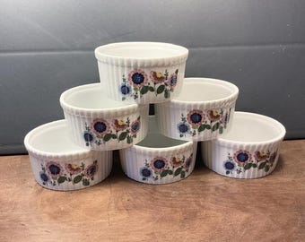May include: Set of six white ceramic ramekins with a floral design. The ramekins are stacked in a pyramid shape. The flowers are pink, blue, and yellow.