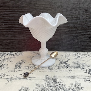 Puede incluir: Plato de caramelo de vidrio de leche blanco con un borde festoneado y un patrón de diamante. El plato está sobre un pedestal y tiene una pequeña cuchara al lado.